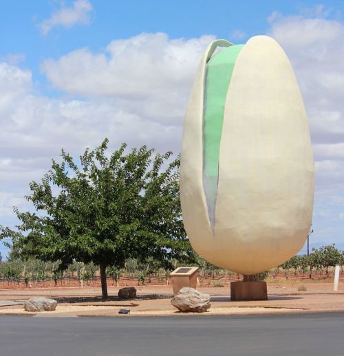 A large, sculptural pistachio nut stands on a pedestal, surrounded by trees and a blue sky with clouds.