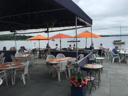 Outdoor dining area by the water, featuring orange umbrellas, tables, and patrons enjoying the view.