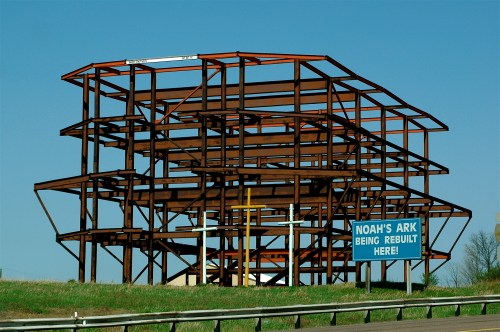 Steel framework of a building under construction with a sign reading "Noah's Ark Being Rebuilt Here!" against a clear sky.