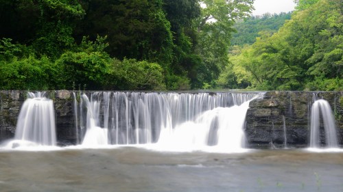 A serene waterfall cascading over a rocky ledge, surrounded by lush green trees and a calm river.