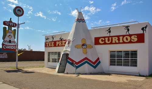 A colorful roadside shop shaped like a teepee, featuring signs for "Curios" and "Tee Pee Curios" against a blue sky.