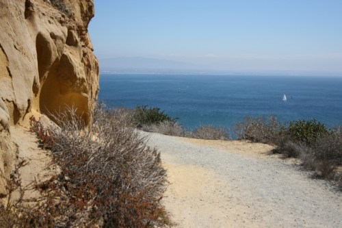 A scenic coastal path with rocky cliffs on one side and a view of the ocean and a sailboat in the distance.