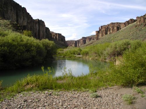 A serene river flows through a lush green valley surrounded by towering cliffs under a blue sky.