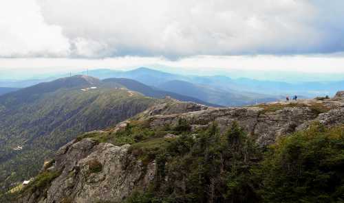 A panoramic view of mountains under a cloudy sky, with hikers on a rocky ledge in the foreground.