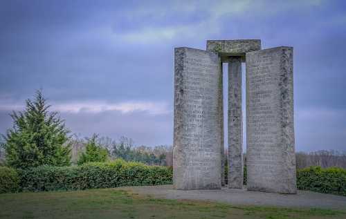 A stone monument with tall pillars inscribed with text, surrounded by greenery under a cloudy sky.