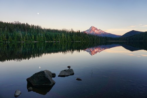 A serene lake reflects a snow-capped mountain and a clear sky, surrounded by lush green trees at dusk.