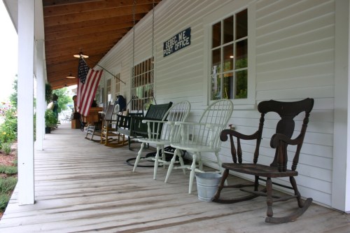 A porch with rocking chairs and an American flag, featuring a sign for a post office.