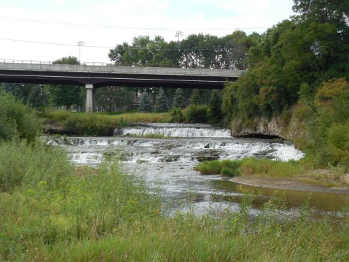 A serene river with cascading waterfalls, framed by lush greenery and a bridge overhead.