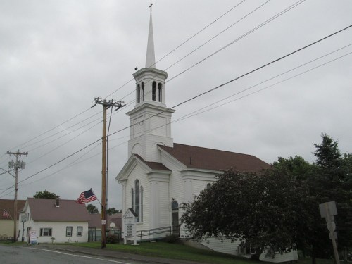 A white church with a tall steeple, surrounded by power lines and trees, under a cloudy sky. An American flag is visible.