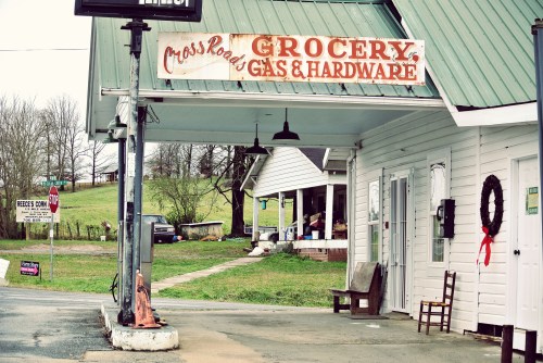 A small grocery store with a sign reading "Grocery, Gas & Hardware," featuring a rustic exterior and nearby greenery.