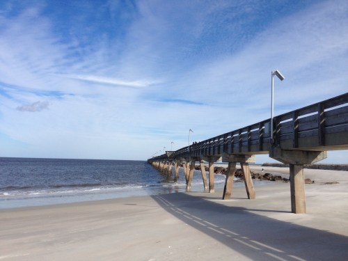 A wooden pier extends over calm waters, with a sandy beach and blue sky in the background.