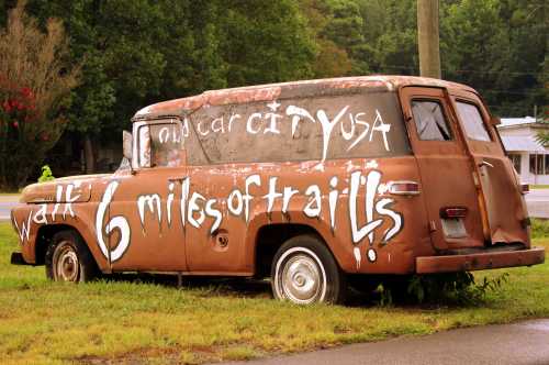 A rusty old truck with graffiti promoting "Walk 6 miles of trails" in a grassy area.