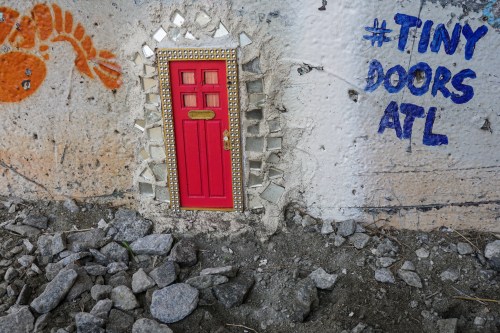 A small red door set into a wall, surrounded by mosaic tiles and graffiti reading "#TINY DOORS ATL."
