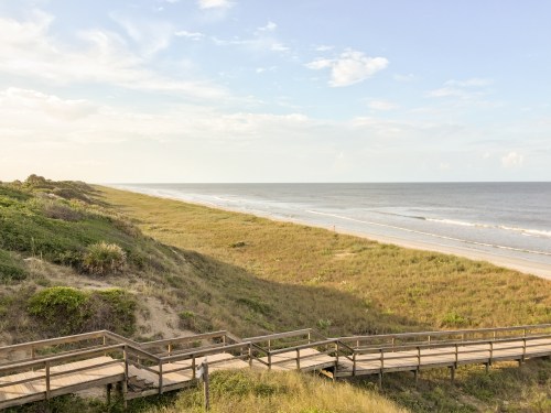 A wooden walkway leads to a serene beach with gentle waves and grassy dunes under a clear sky.