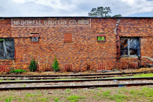 Weathered brick wall of an old building with faded "McDowell Grocery Co." sign and overgrown vegetation near railway tracks.