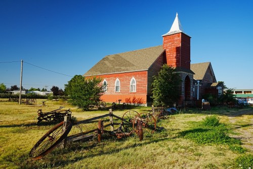 A red church with a steeple surrounded by overgrown grass and old farm equipment in a clear blue sky.