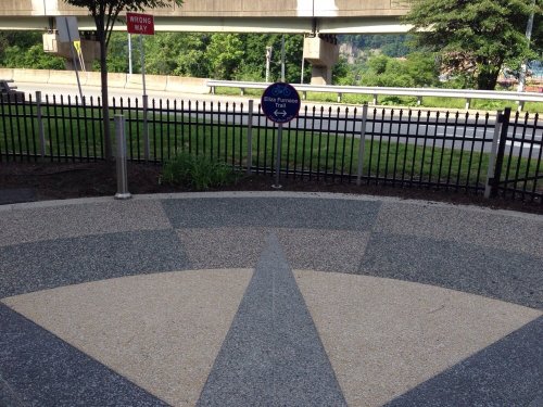 A circular paved area with a directional sign, surrounded by greenery and a fence, near a road under a bridge.