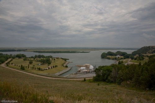 A scenic view of a lake surrounded by greenery and a cloudy sky, with a small dock and boathouse in the foreground.