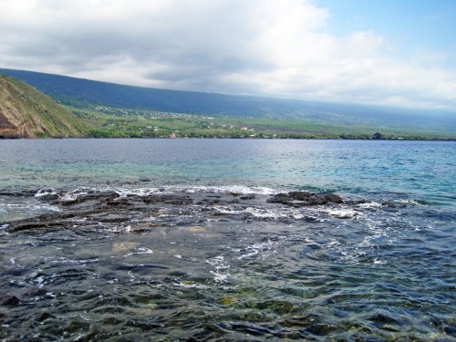 A serene coastal view with rocky shoreline, clear water, and distant green hills under a partly cloudy sky.