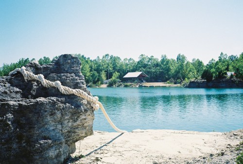 A rocky shoreline with a rope leading to a calm blue lake, surrounded by lush green trees and a distant cabin.