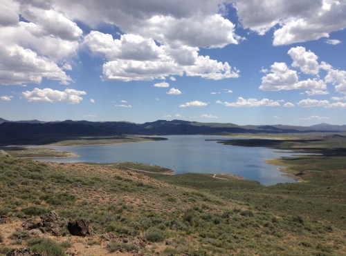 A serene lake surrounded by rolling hills and mountains under a blue sky with fluffy white clouds.