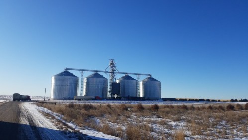 Metal grain silos stand against a clear blue sky, surrounded by snow-covered fields and a gravel road.