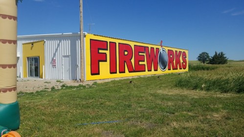A large yellow wall with bold red letters spelling "FIREWORKS" next to a grassy field and clear blue sky.