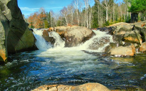 A serene waterfall cascades over rocks, surrounded by trees and a clear blue sky.