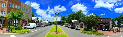 A vibrant street scene with shops, parked cars, and a clear blue sky, showcasing a lively urban atmosphere.