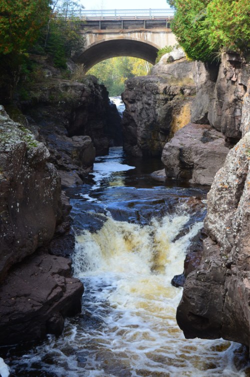 A scenic view of a river cascading through rocky cliffs, with a bridge arching overhead amidst lush greenery.