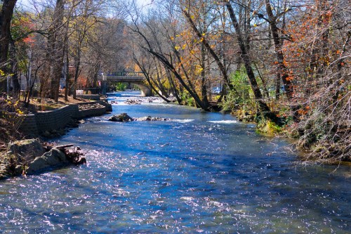 A serene river flows through a wooded area, with trees displaying autumn colors and a bridge in the background.