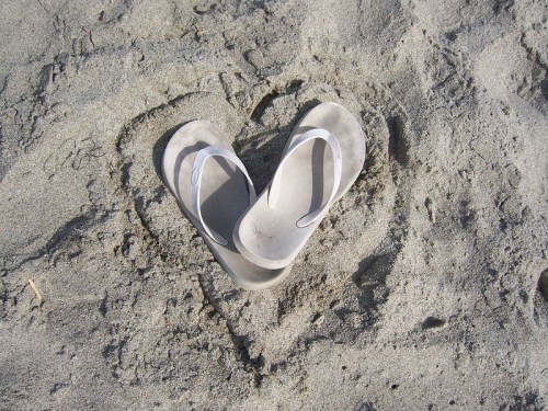 A pair of flip-flops arranged in a heart shape on sandy beach.