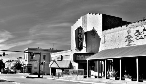Black and white image of the Rose Theater building with vintage signage, surrounded by small shops and a clear sky.