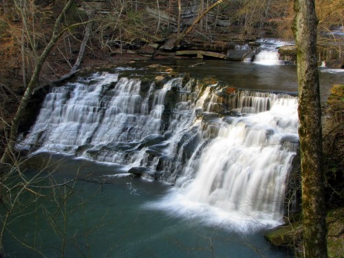 A serene waterfall cascades over rocky ledges, surrounded by trees and a tranquil river.