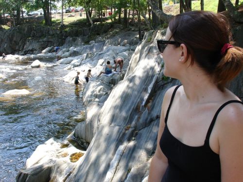 A woman in sunglasses sits by a river, watching people play on the rocks and in the water. Trees line the background.