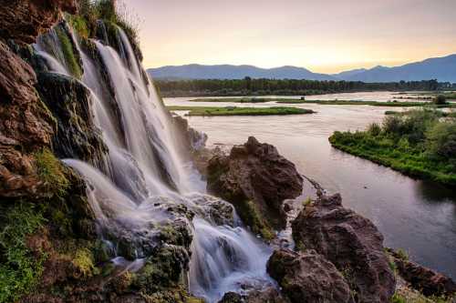 A serene waterfall cascades over rocky cliffs into a calm river, surrounded by lush greenery and distant mountains at sunset.