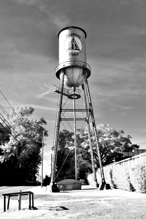 Black and white image of a tall water tower with a triangular logo, surrounded by trees and power lines.