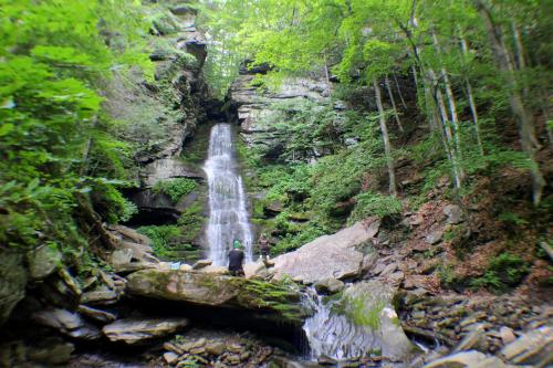 A serene waterfall cascades down rocky cliffs, surrounded by lush greenery and trees, with two people exploring nearby.