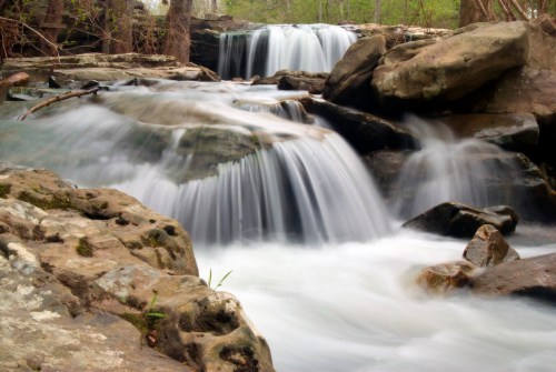 A serene waterfall cascades over rocky terrain, surrounded by lush greenery and flowing water.