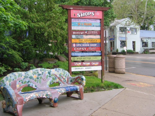 Colorful signpost listing various shops, with a decorative bench in the foreground and trees in the background.