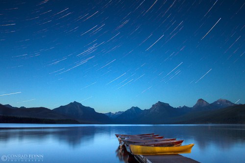 A serene lake at night with star trails above, featuring a dock with colorful boats and mountains in the background.