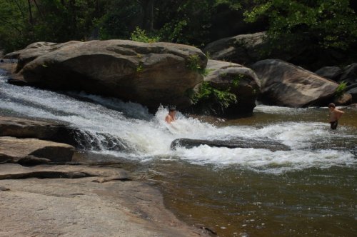 A person splashes in a river near large rocks, surrounded by lush greenery on a sunny day.