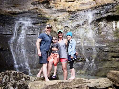 A family of four stands on rocks by a waterfall, smiling and enjoying a day outdoors.