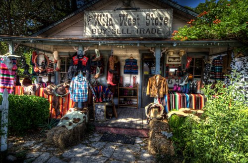 A rustic storefront displaying colorful clothing and accessories, with a sign reading "Wild West Store: Buy, Sell, Trade."