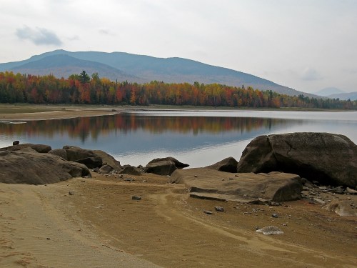 A serene lake surrounded by autumn foliage, with mountains in the background and rocks in the foreground.
