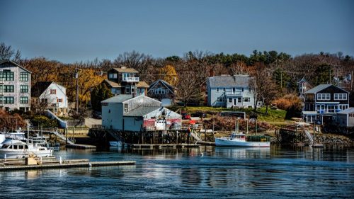 A scenic waterfront view featuring boats, houses, and trees along a calm river on a clear day.