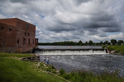 A brick building beside a river with a waterfall, under a cloudy sky, and two people standing near the water's edge.