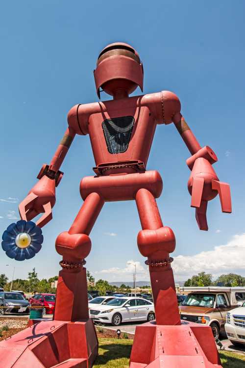 A large red robot sculpture stands in a parking lot, holding a flower in one hand against a clear blue sky.