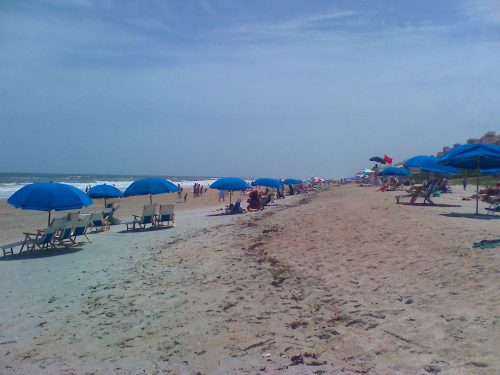 A sandy beach scene with blue umbrellas, lounge chairs, and people enjoying the sun near the ocean.