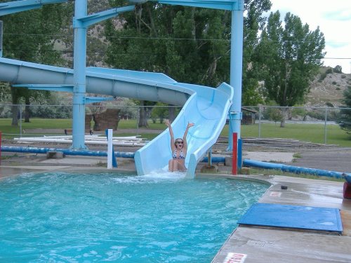 A person joyfully sliding down a water slide into a pool on a sunny day, surrounded by trees and a fence.